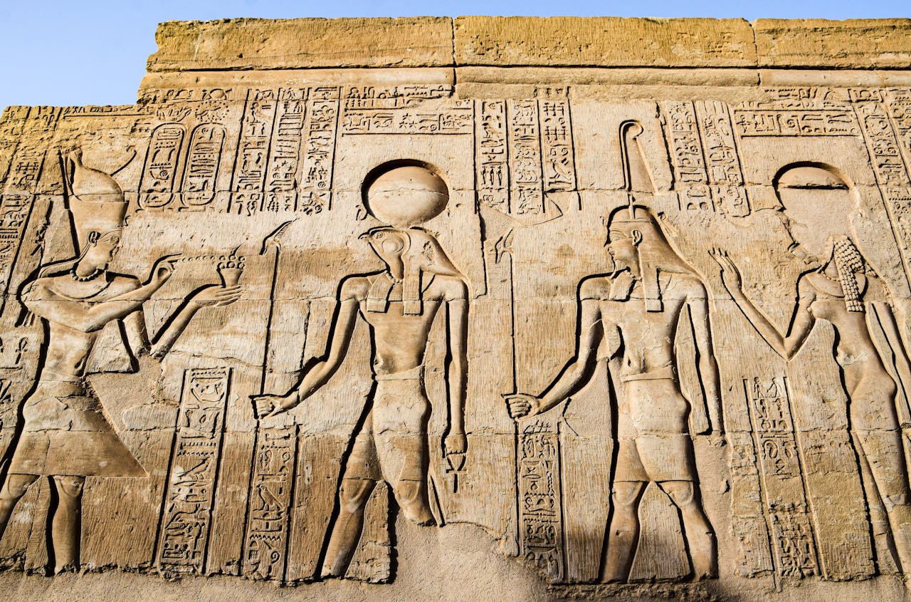 Detailed carvings of ancient Egyptian deities and hieroglyphs on a temple wall in bright daylight.